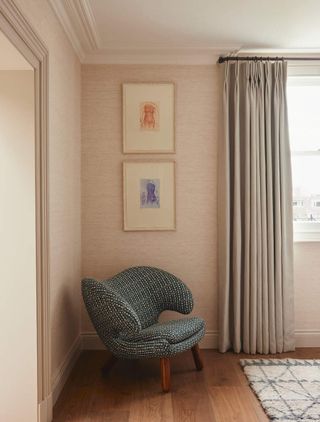 Image of the corner of a pink living room. The walls are a blush pink and the ceiling is white with crown molding. There is a dark green, modern accent chair in the corner and two pictures hanging above it. There is a window partially in frame with light gray curtains that are open.