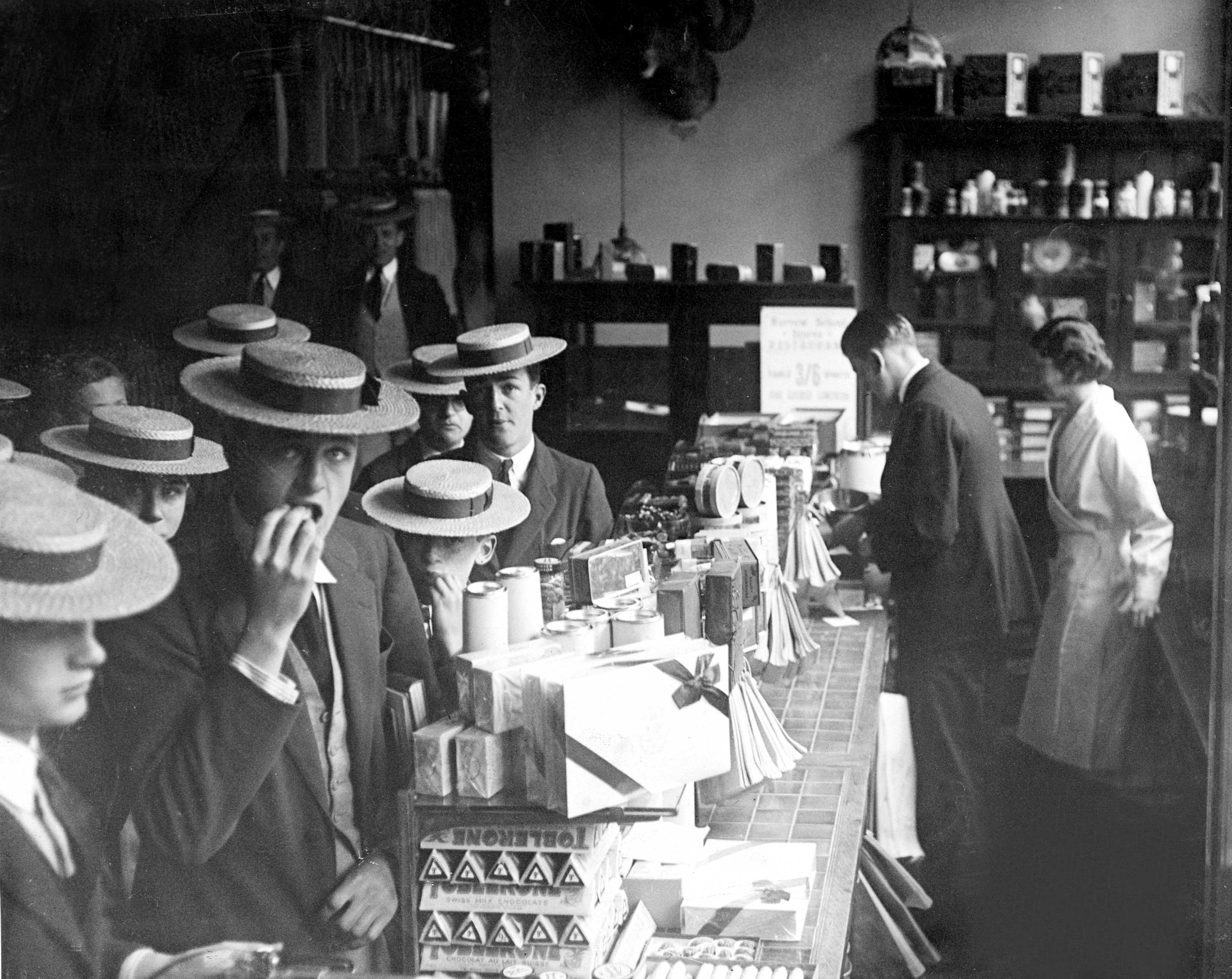 Boys in straw boater hats crowd along a shop counter stacked with Toblerone and boxes of sweets, one of them looking at the camera while eating.