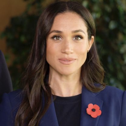 a close up of Meghan Markle wearing a navy blazer with a black T shirt and a flower lapel pin