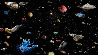 Plastic oceanic debris appears colorfully in sharp contrast against the pitch black of the ocean, items include a toothpaste tube and other recognizable waste