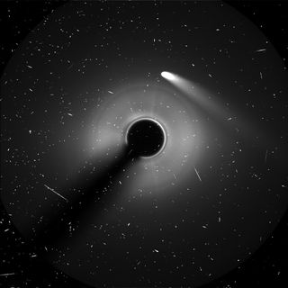 the white streak of a comet stretches downward right from the center, near a black circle surrounded in hazy white, against the starry black of space.