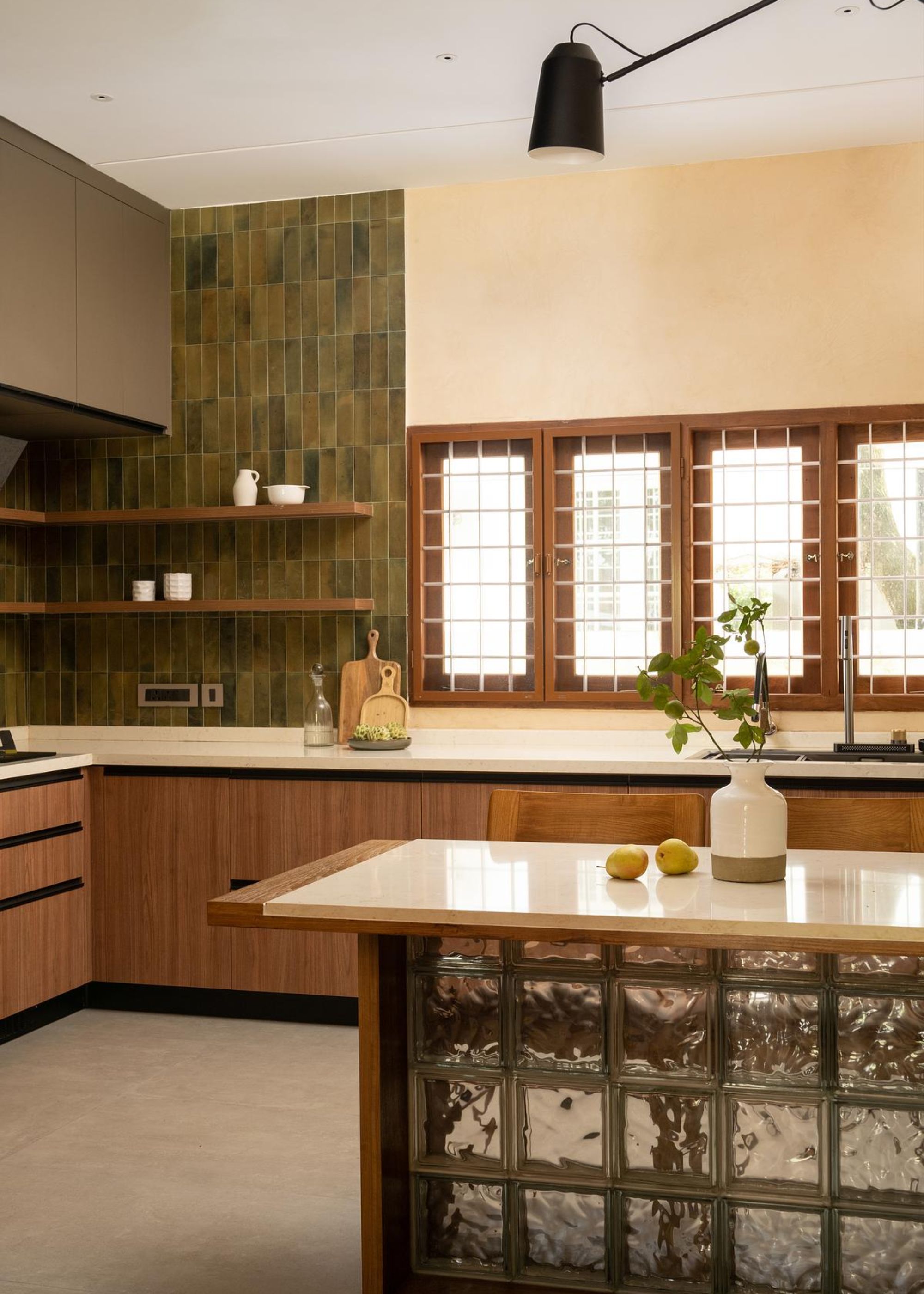 A kitchen with a glass fluted island and a green tiled backsplash with open shelving on top of it as well as wooden framed windows. On the island is a vase with a plant as well as some fruits on top.