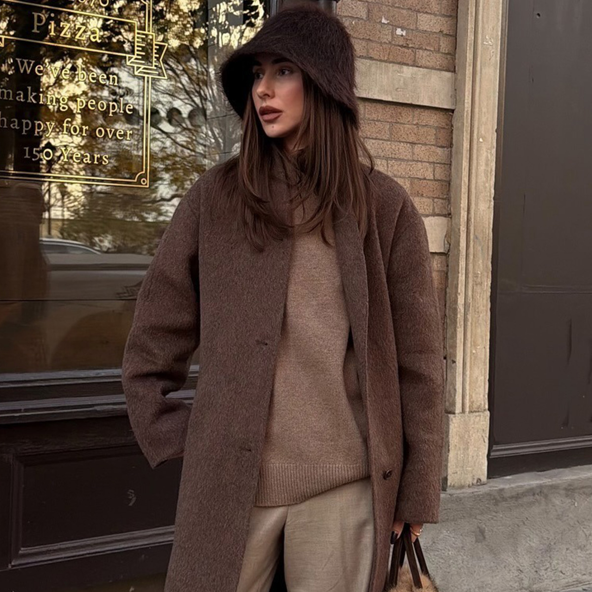 Madina wears a fur hat, brown wool coat, brown sweater, and tan pants.