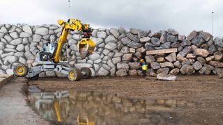 a yellow excavator holds a large boulder, part of its effort to build a rock wall