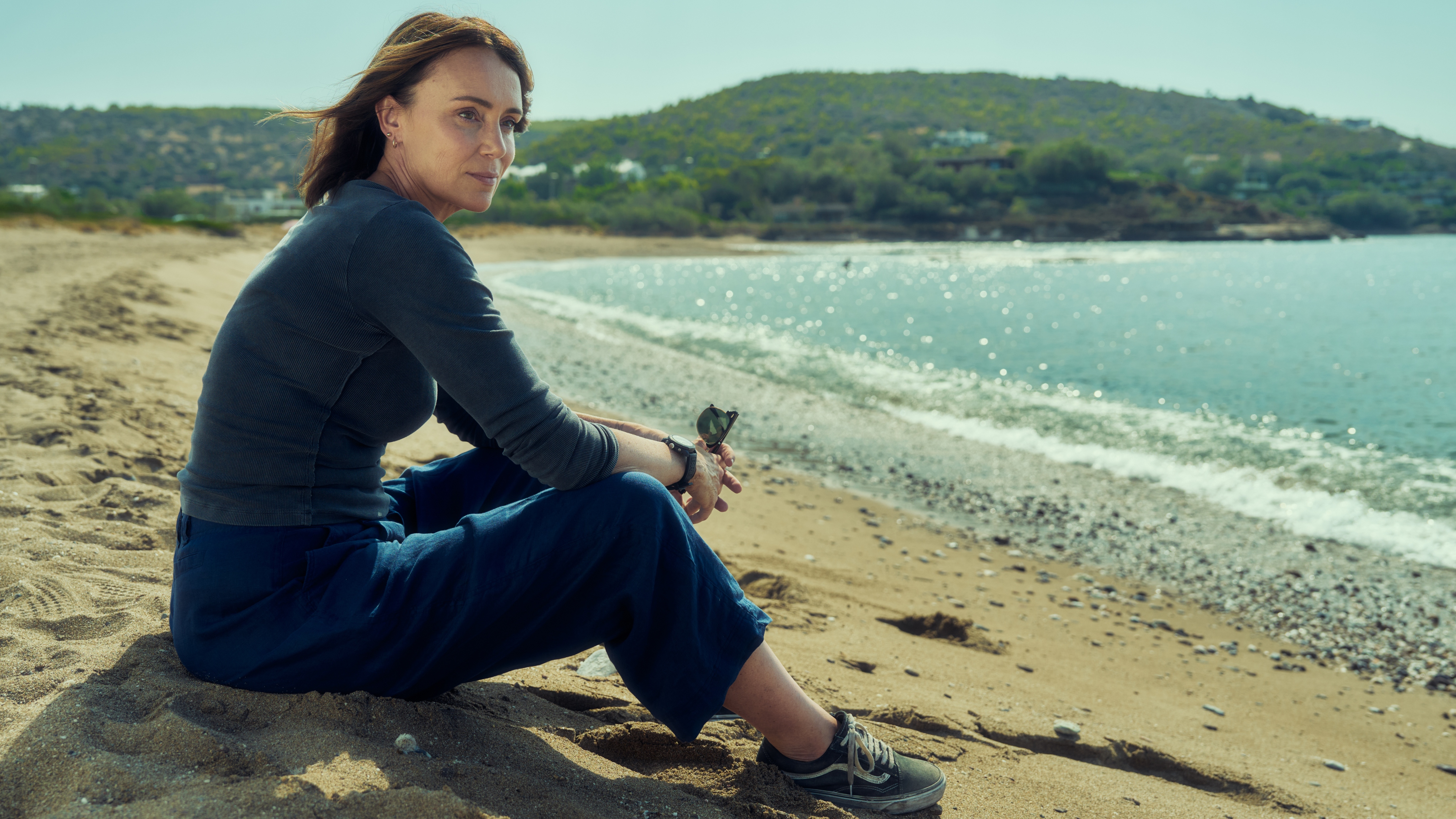 Julie from The Assassin sitting on the beach looking out to sea in a blue top and trousers.