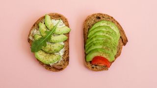 Two slices of avocado on toast, one with rocket leaf, on pastel pink background
