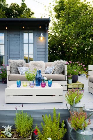 a blue wood garden room behind a white patio with white garden furniture, decorated with flower pots and watering can planters