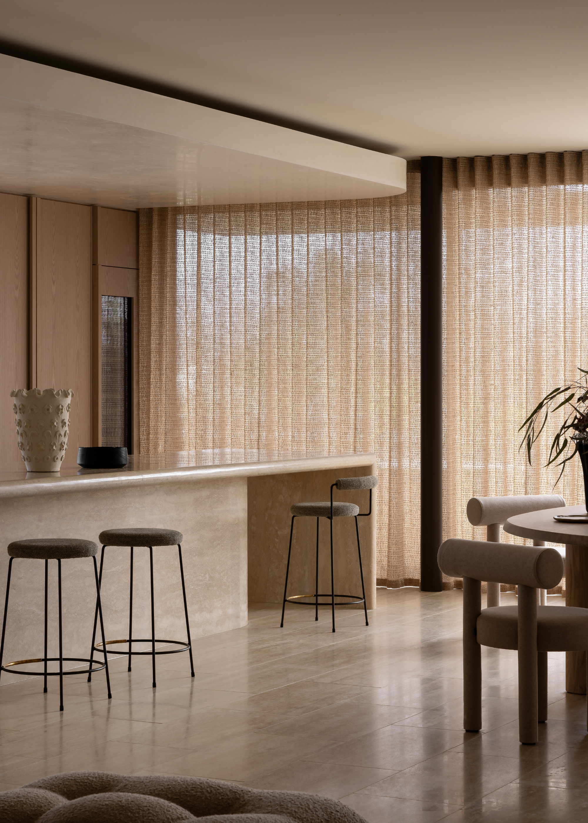 An open kitchen and dining room with black and gray bar stools, a textured vase, a round table with geometric chairs, and sheer textured curtains