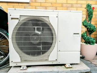 A heat pump outside a brick house with a fake plastic plant beside it