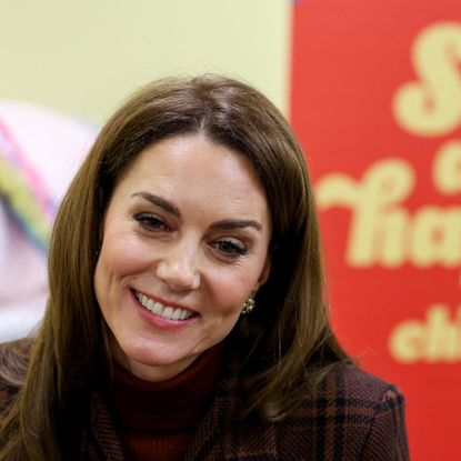 A shoulder up photo of Kate Middleton wearing a burgundy plaid coat sitting in front of a red sign saying safe and happy childhood in yellow letters