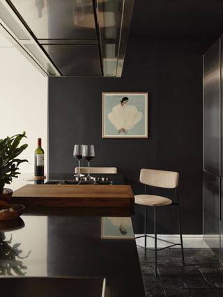 Image of a home bar area that is painted in a dark gray-blue color. There is a light blue artwork hanging on the wall, and two white bar stools set up around a couple glasses of wine.