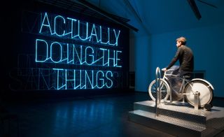 Pic 1 of 3 - man on a bicycle in front of a neon sign reading "ACTUALLY DOING THE THINGS"