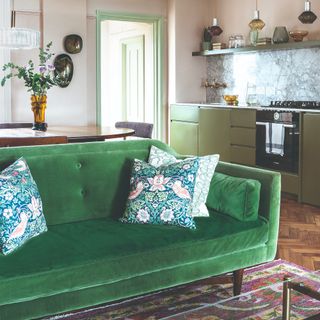 A living room with an emerald green velvet sofa, connected to a kitchen with green cabinets