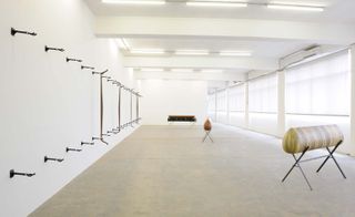 Long white room, ceiling lights, wall of windows right wall, rows of black walls hooks on left, grey marble effect floor, three wooden art pieces on black stands placed around the room