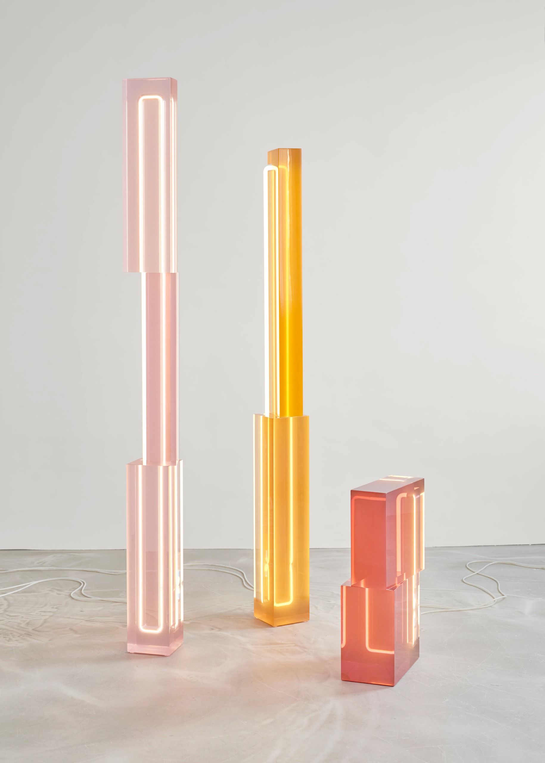 Totem lights made of semi-transparent resin in pink, yellow, and orange, of decreasing height, styled in an otherwise empty gallery.