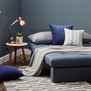 a contemporary blue single bed with blue and white/grey cushions, a grey blanket, a grey and white patterned, and a white marble side table with rose gold lamp on it
