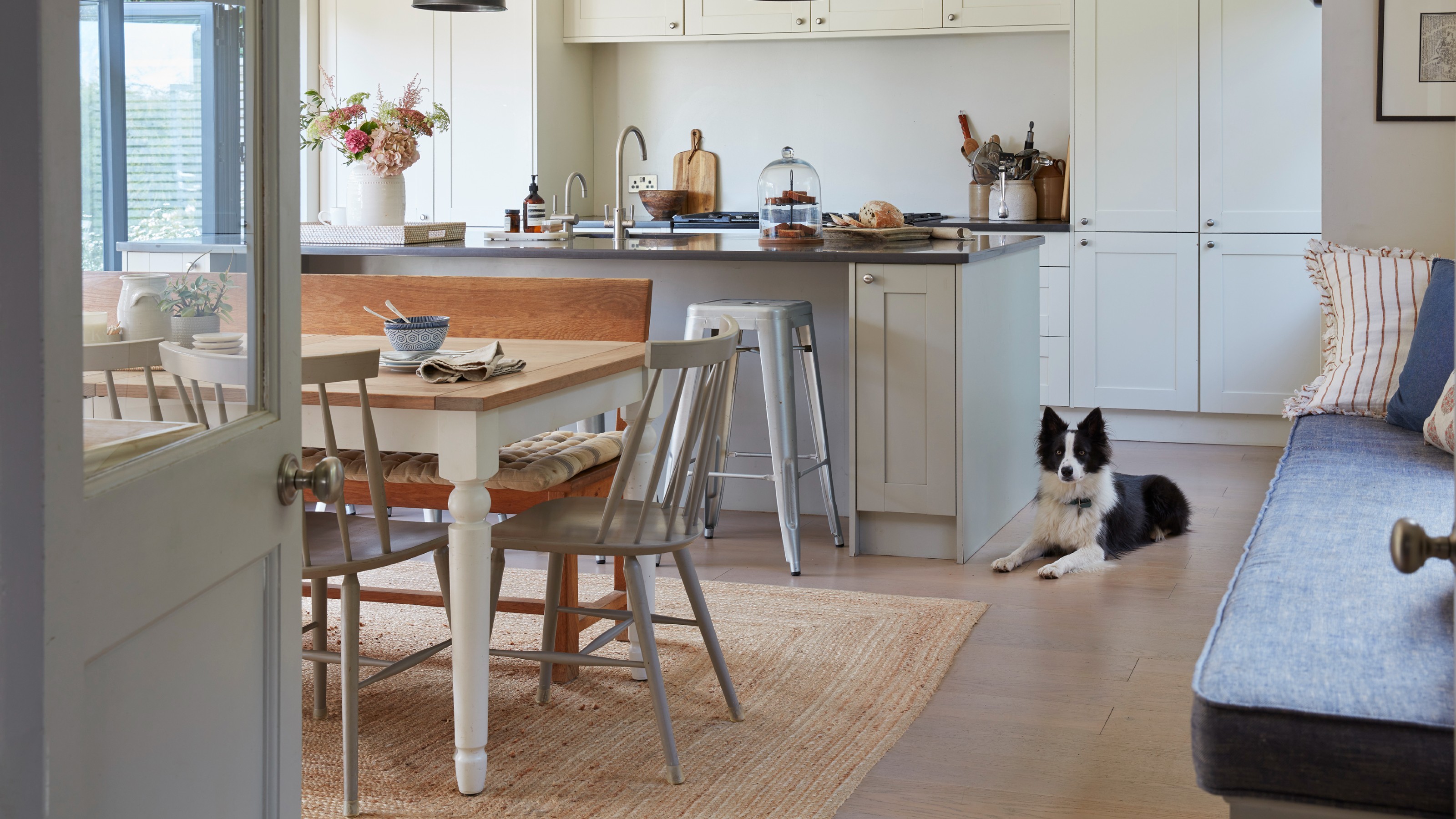 Neutral kitchen diner with jute rug beneath dining table, with a black and white dog and cushioned window seat