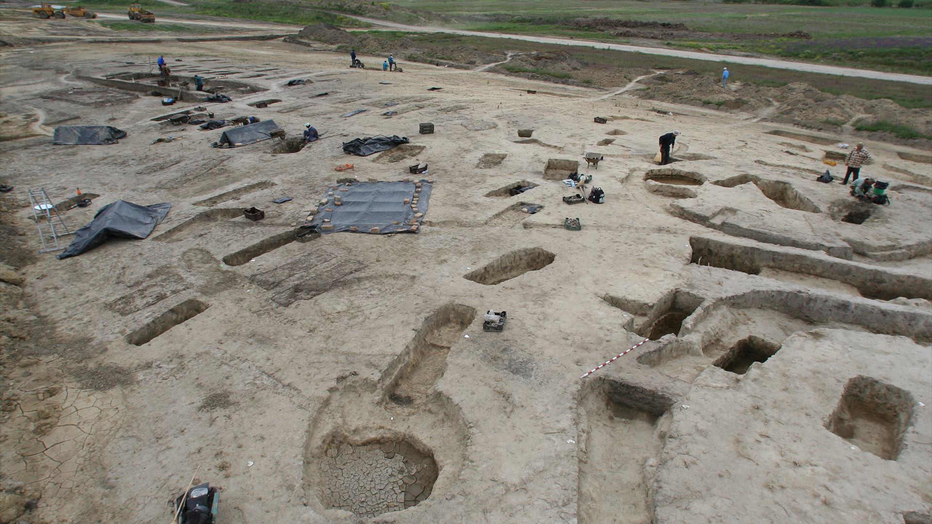 landscape photo of the excavation site, showing holes of different shapes dug up across the field of packed dirt