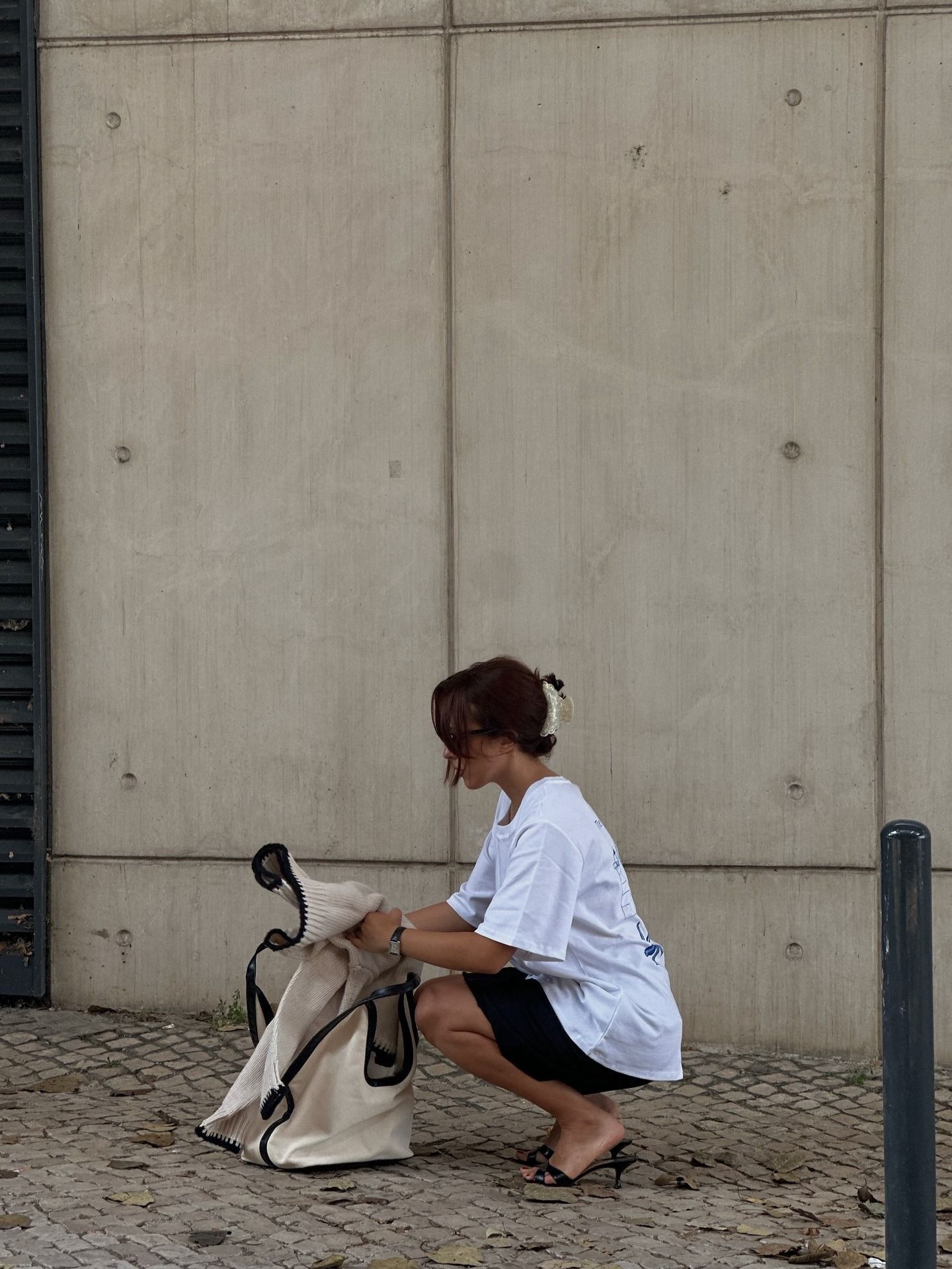 Influencer @deborabrosa crouches down outside, putting a jumper into her bag. She wears a white boxy t-shirt with black shorts and black low-heeled mules.