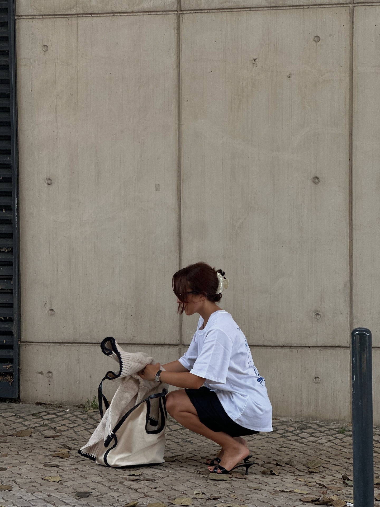 Influencer @deborabrosa crouches down outside, putting a jumper into her bag. She wears a white boxy t-shirt with black shorts and black low-heeled mules.