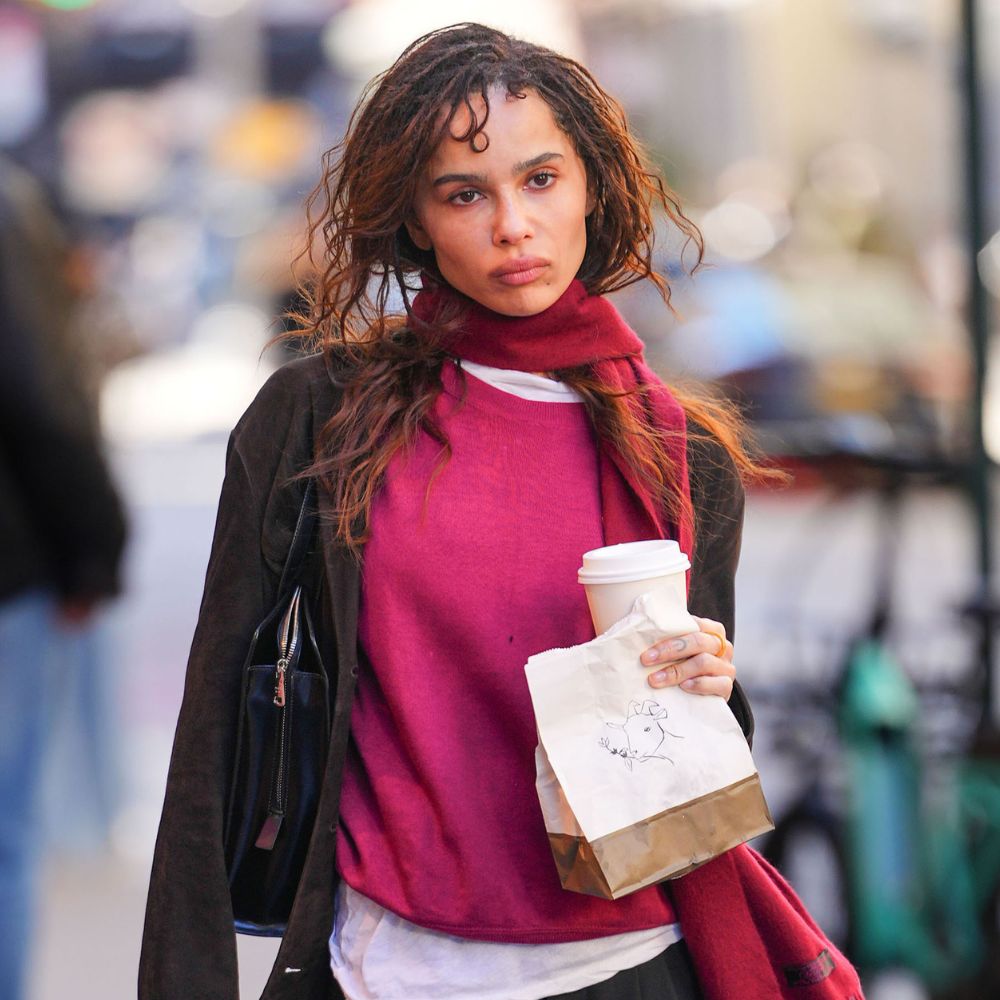 Zoe Kravitz walks down the street in New York wearing black trousers with black trainers, a black jacket and a raspberry jumper and scarf.
