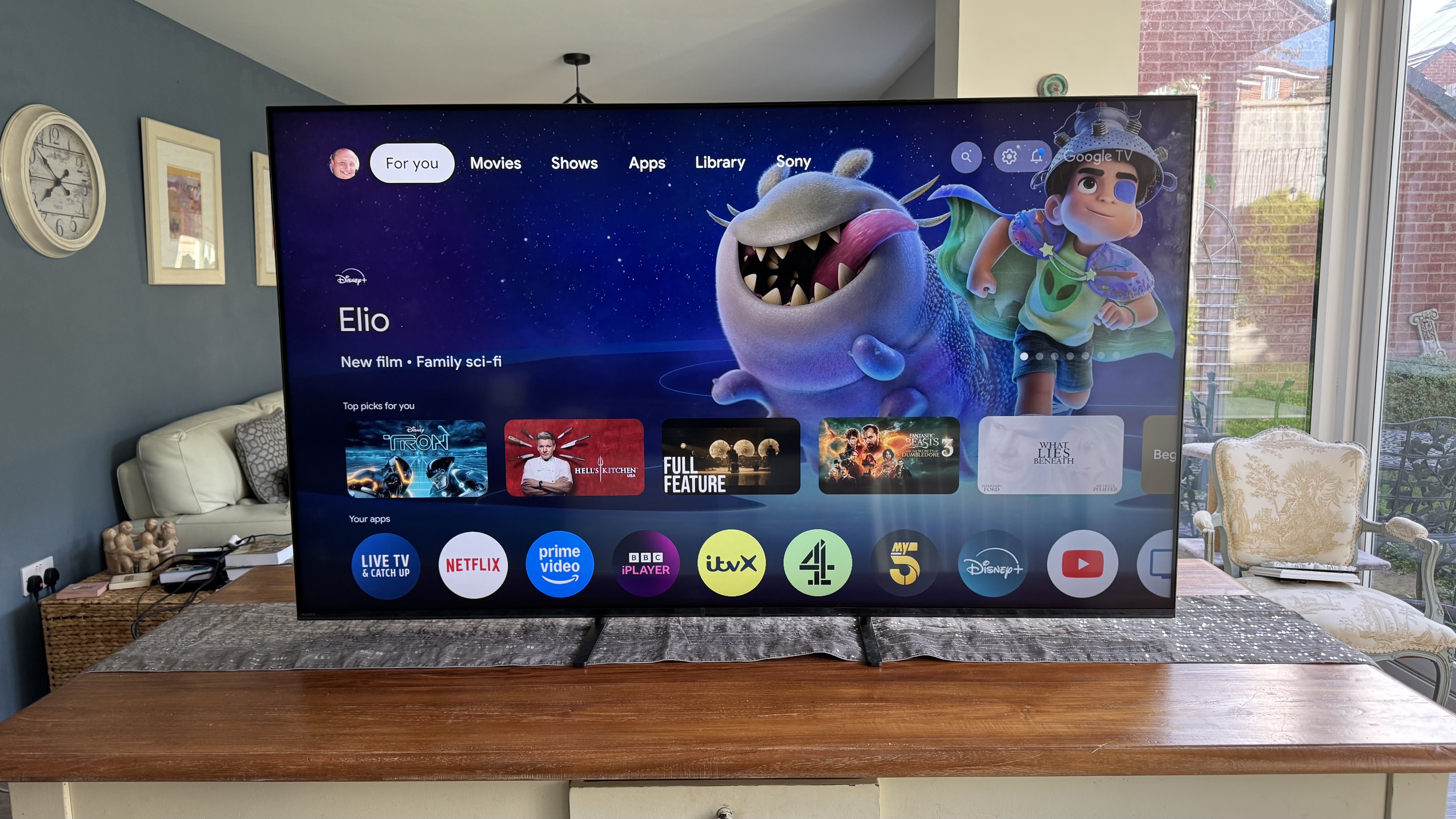 The Sony 65 Bravia 5 TV showing the home screen from a distance, positioned on a wooden cabinet