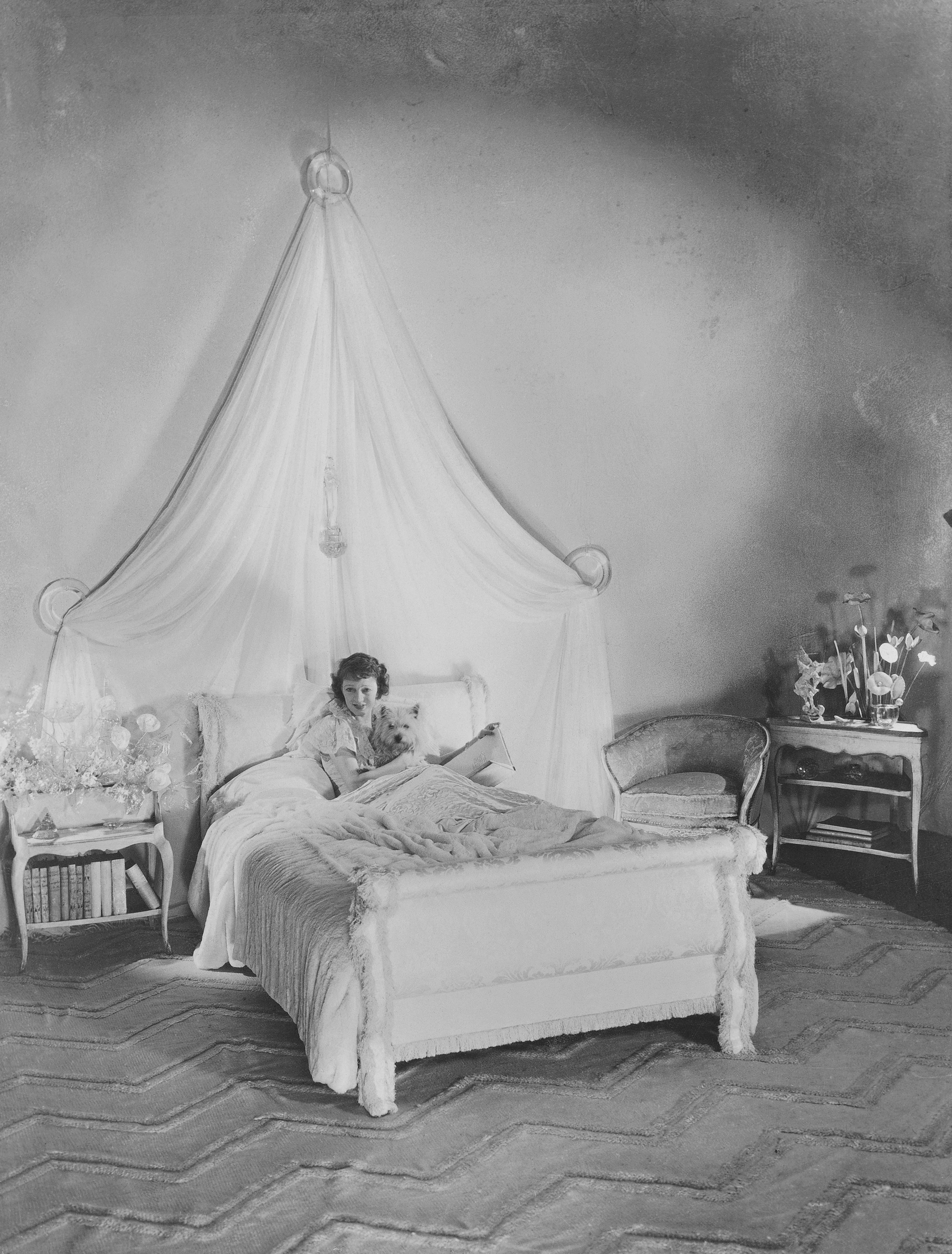 A woman reclines in an ornate white bedroom with a gauze canopy, holding a small shaggy white dog and a book on the bed.