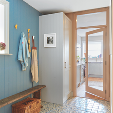 Blue hallway with patterned tiled floor, bench and storage hooks leading into a small kitchen.