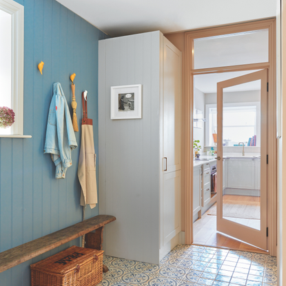 Blue hallway with patterned tiled floor, bench and storage hooks leading into a small kitchen.