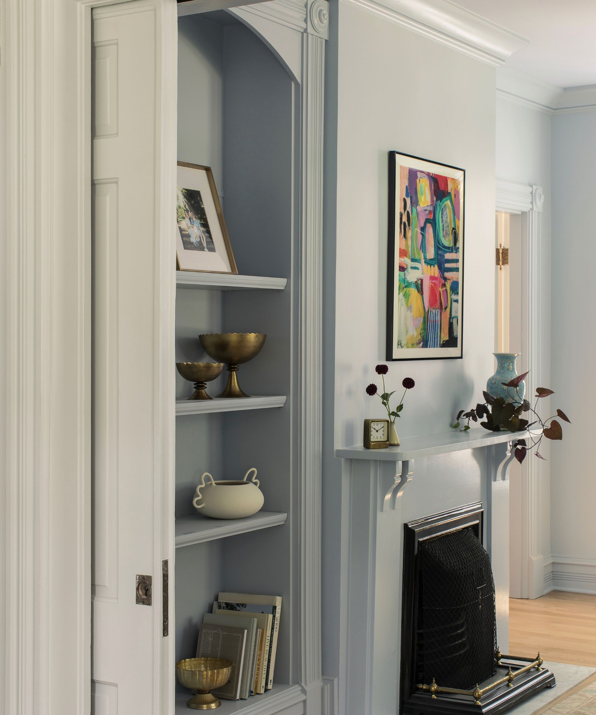 Living room colour drenched in pale blue, a large fireplace and built-in alcove with bookshelves also painted pale blue, decorated with books and decor, and a large abstract print above the mantel.