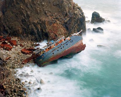 A photo of a rusted ship turned on its side surrounded by rocks and the sea.