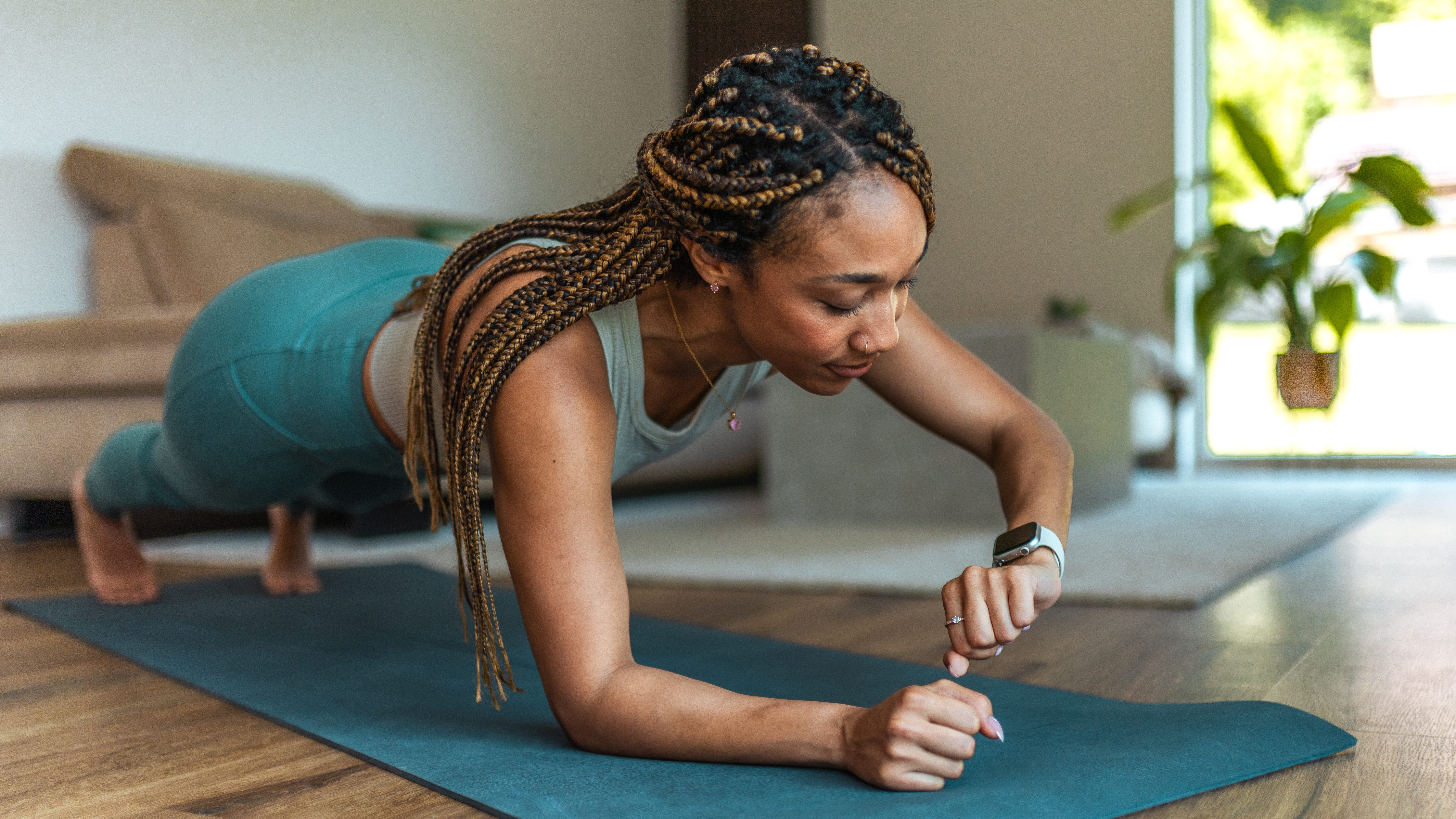 A woman holds a plank on an exercise mat and looks at her fitness tracker. She is resting on her toes and her right forearm, with her body held in a straight line.