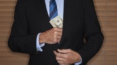 A man wearing a suit slips a stack of cash into his inner breast pocket.