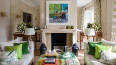 A cream and green living room with a mirrored wall, large mantelpiece with colorful artwork above, two matching cream couches with green cushions and a green coffee table between.