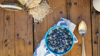 Oats in a glass jar scattered onto wooden table top, next to bowl of overnight oats with blueberries on top