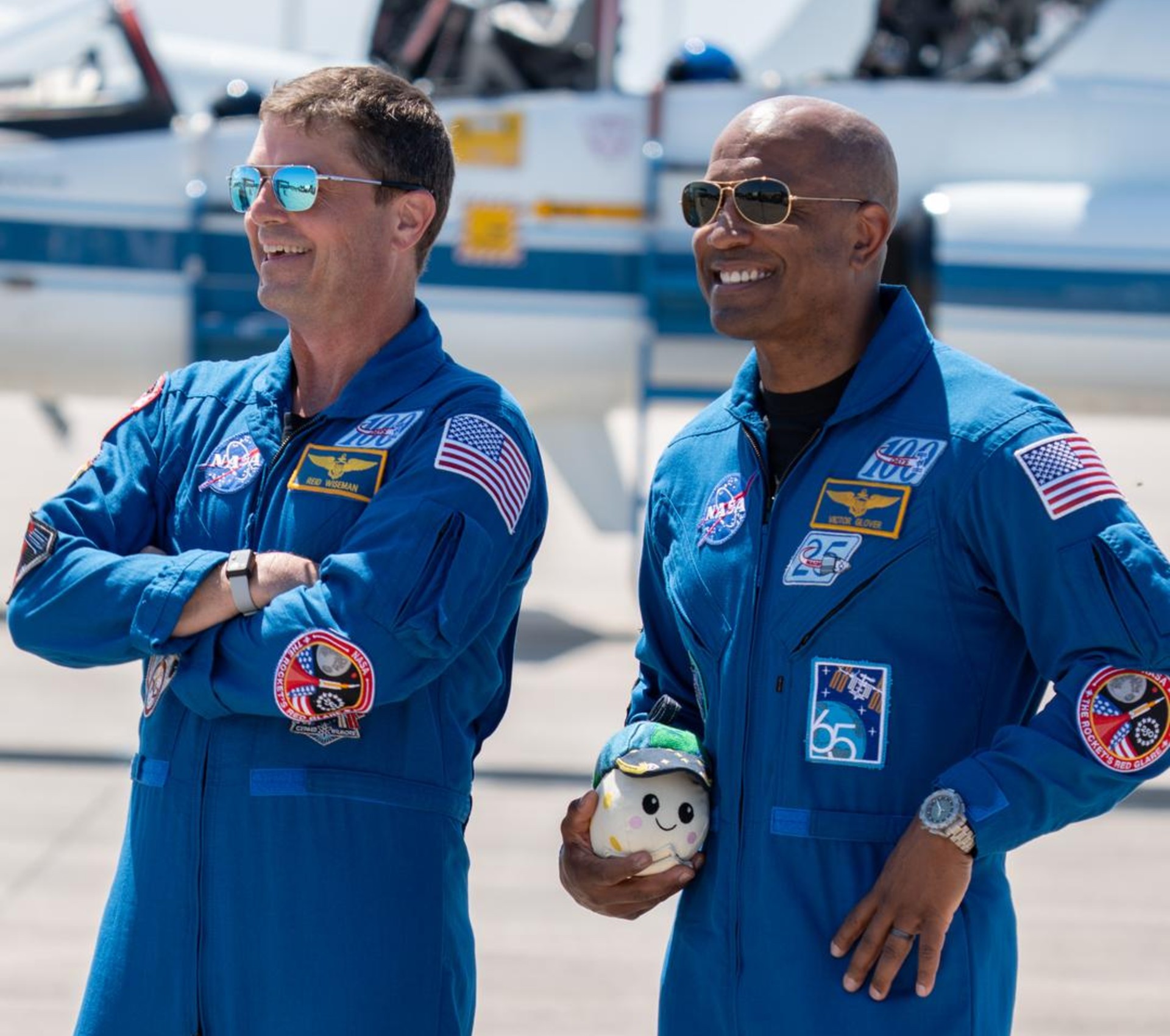 two people in blue flight suits and sunglasses smile on an airplane tarmac. one of them is carrying a small stuffed moon with a smiley face