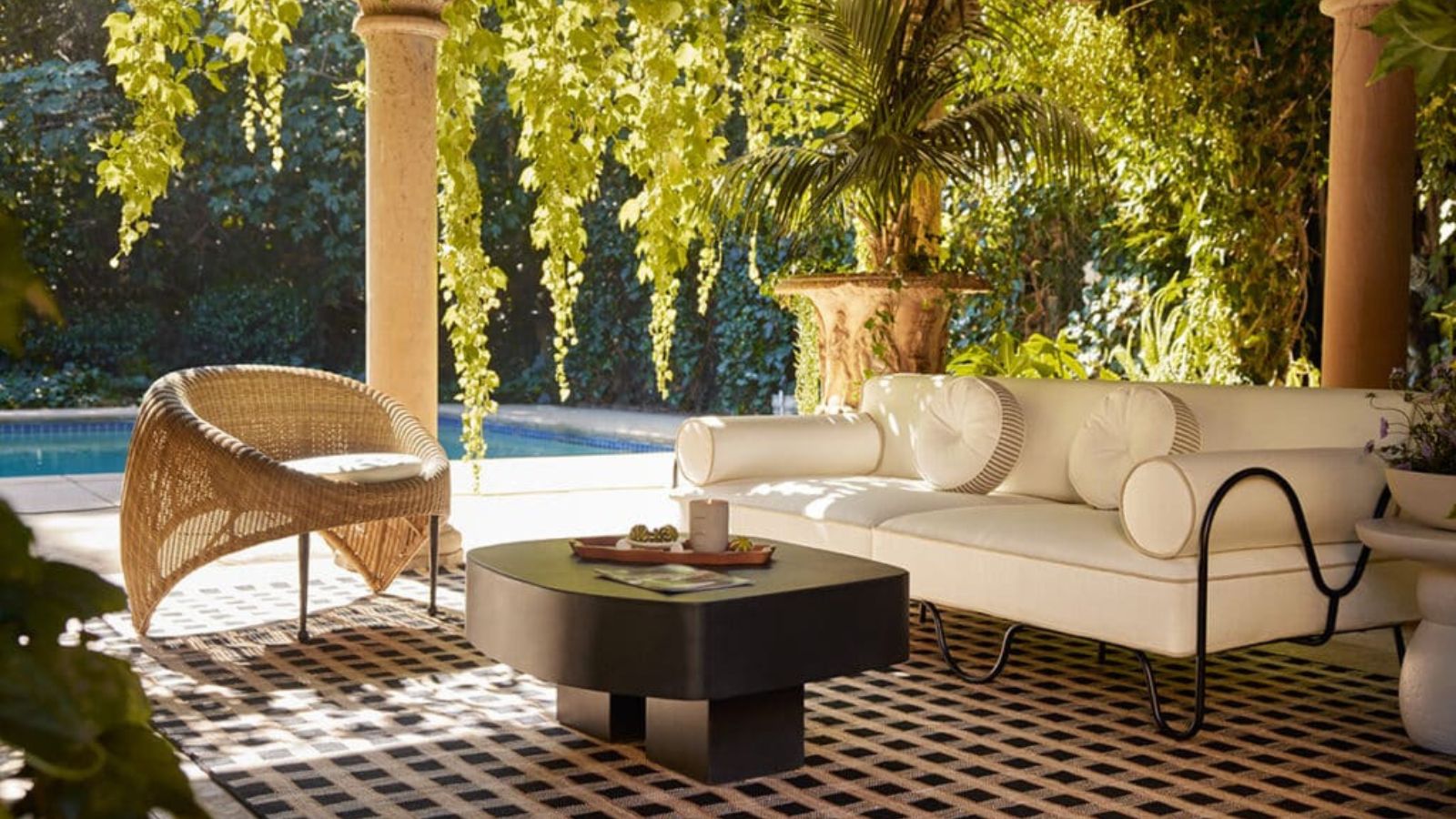 A sofa and an accent chair around a black coffee table on a black and white tiled floor against lush greenery.