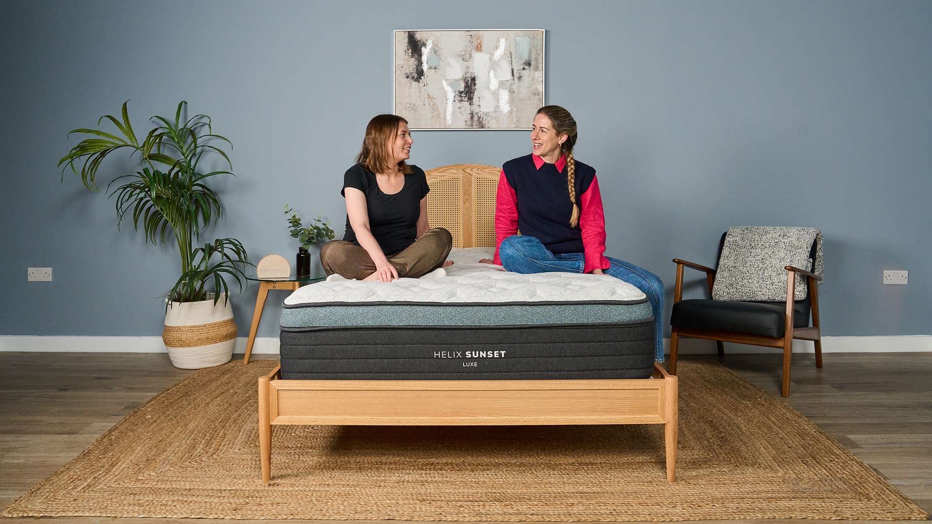 Two women sit on the Helix Sunset Luxe mattress on a bed frame in the Tom's Guide sleep lab