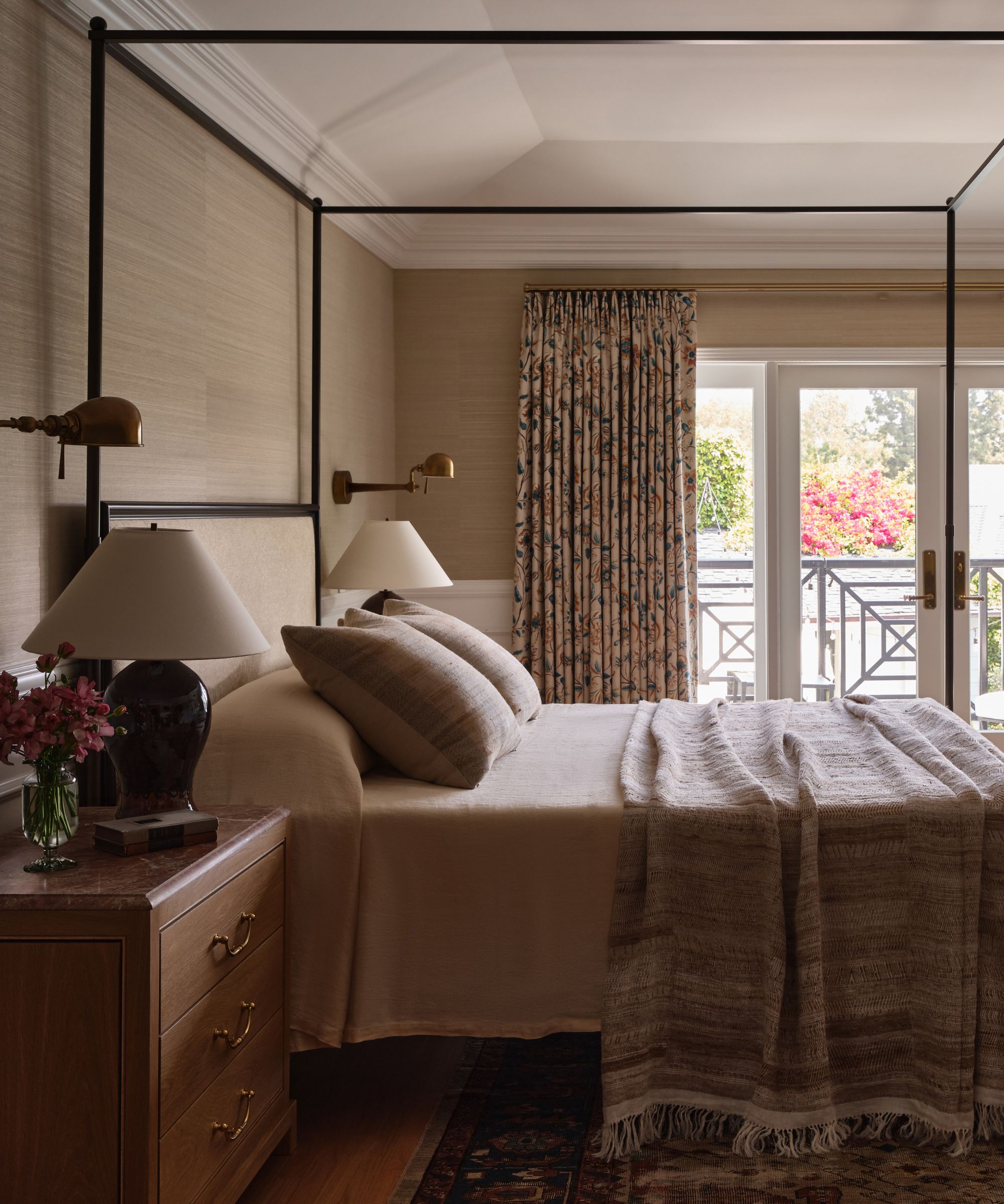 Luxurious bedroom with a four-poster bed, neutral textured wallpaper, patterned curtains, a fluffy armchair, and French doors opening to a balcony garden