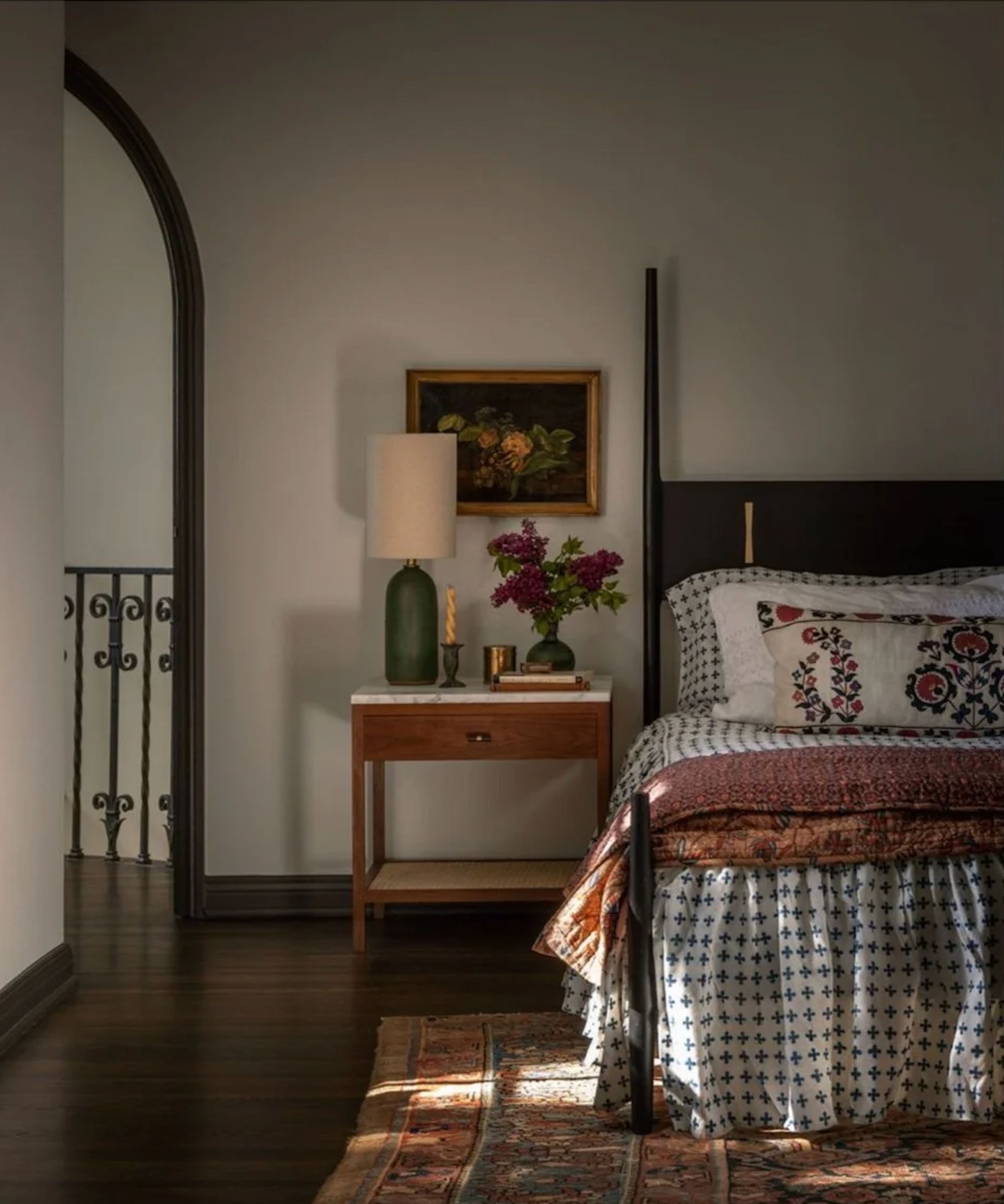 A vertical shot of a cozy, dark-toned bedroom. The bed has patterned white and blue linens and a red floral pillow. To the left, a walnut nightstand with a white marble top holds a tall green lamp and a vase of lilacs. Sunlight streaks across a vintage Persian rug on the dark hardwood floor.