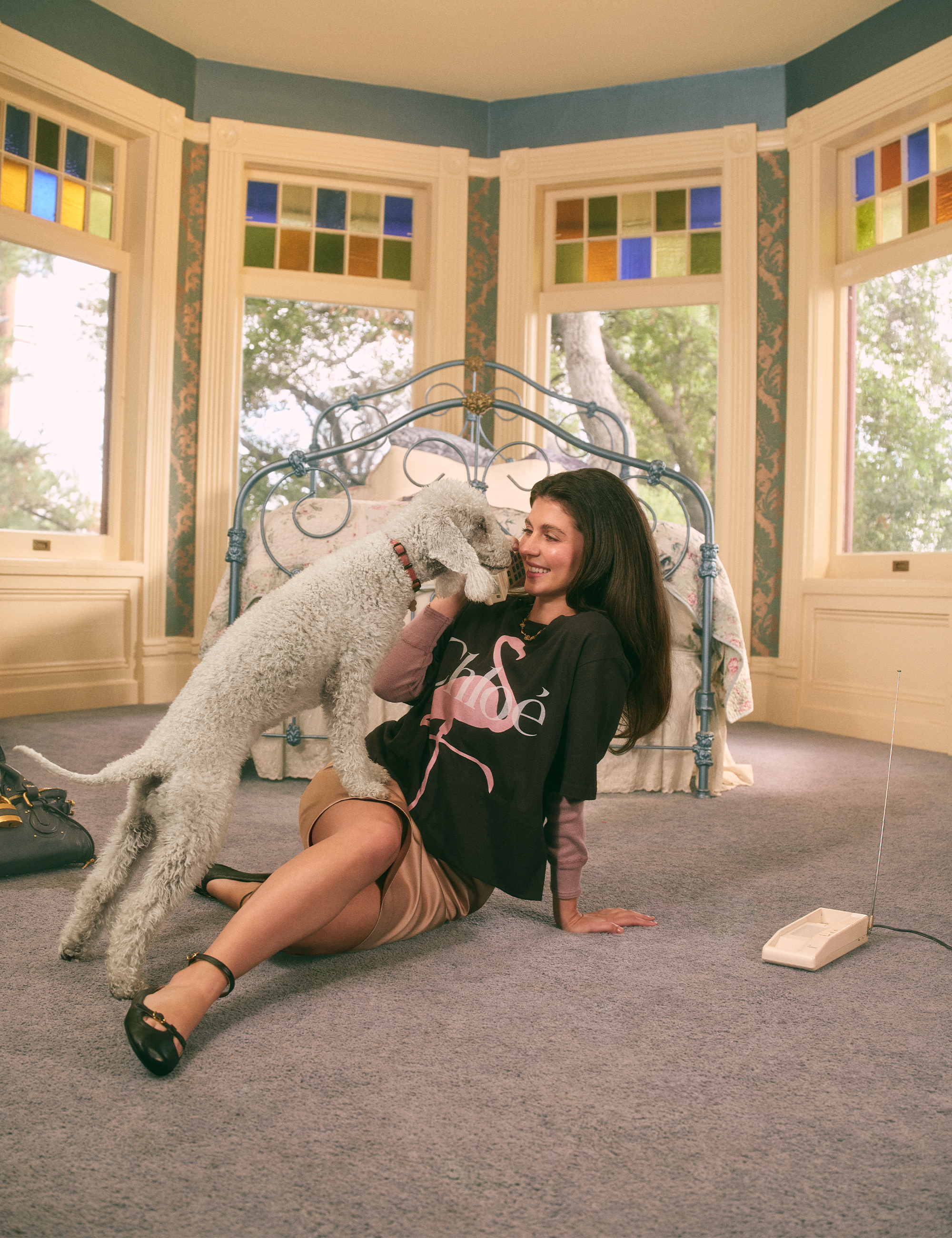 Emily Bader is photographed for Who What Wear's In Focus feature sitting on the floor of a bedroom playing with a poodle who is licking her face. She is wearing a Chloe t-shirt with a pink flamingo on the front and a pink silk skirt.