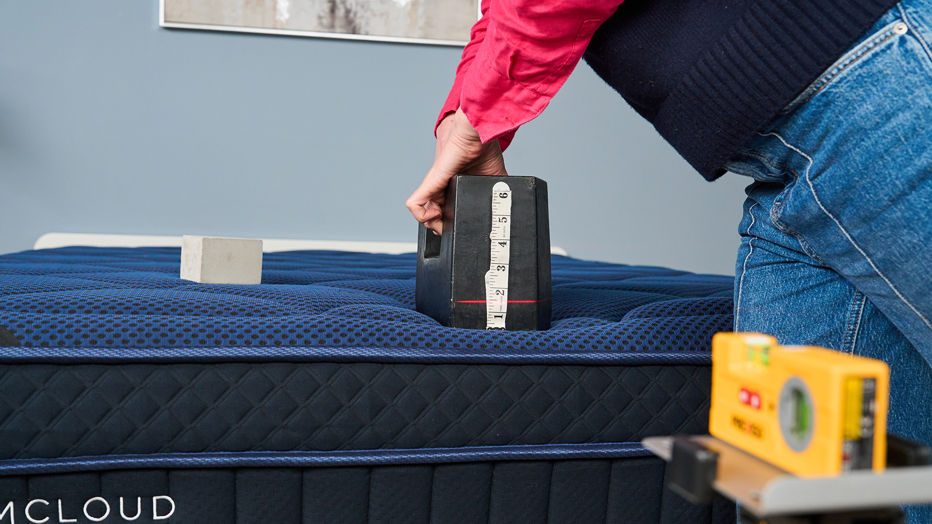 The image shows a mattress tester wearing a pink shirt and blue sweater vest place a 25kg weight on the base and side of a mattress to determine its edge support