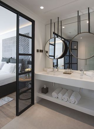 Interior view of the sleeping and bathroom areas in a room at The Serras featuring wood flooring and white walls. Behind the bed there is a section with a black and white pattern, the bed has black and white pillows and linen and there is a round side table with a clear vase and flowers beside it. In the bathroom area there are spotlights, a large wall mirror, a smaller round mirror suspended from the ceiling, two sinks, a tissue box, toiletries and white rolled towels