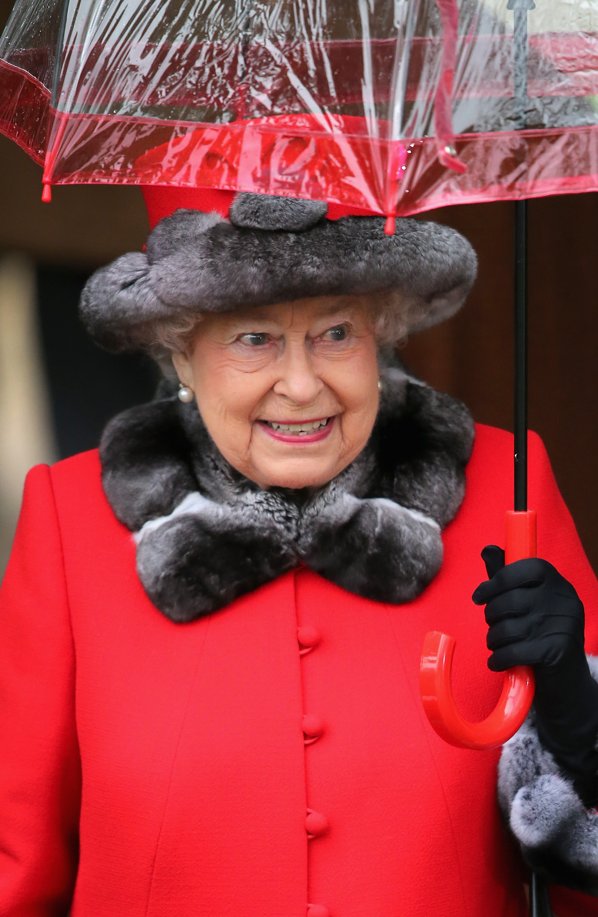 Queen Elizabeth wearing a red hat and coat with fur trim holding a clear umbrella