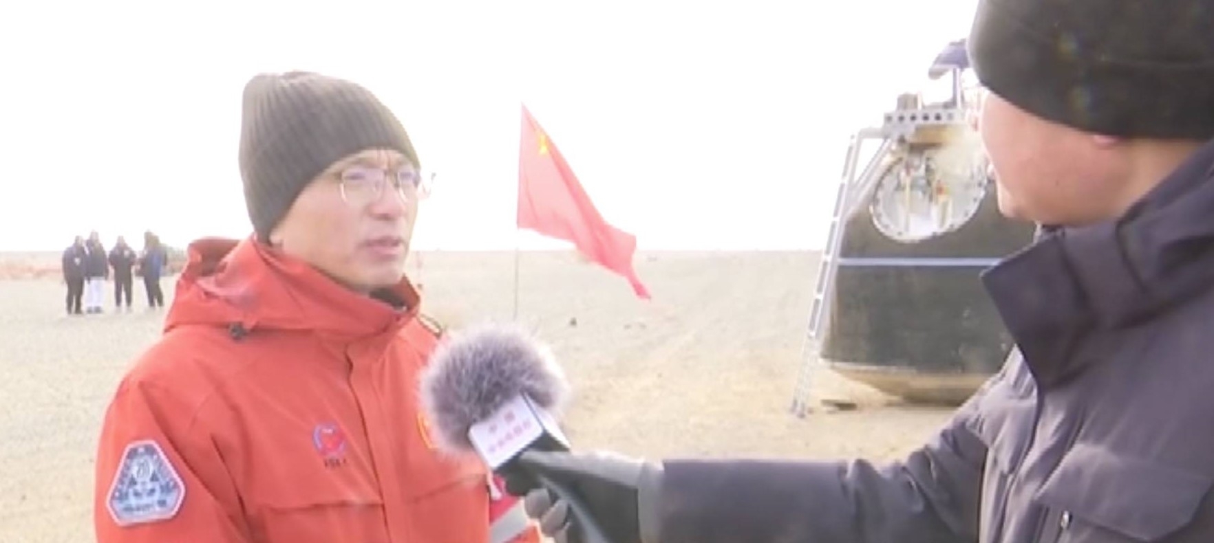 A man wearing a red jacket and black beanie with glasses answers a person wearing a black jacket holding a microphone