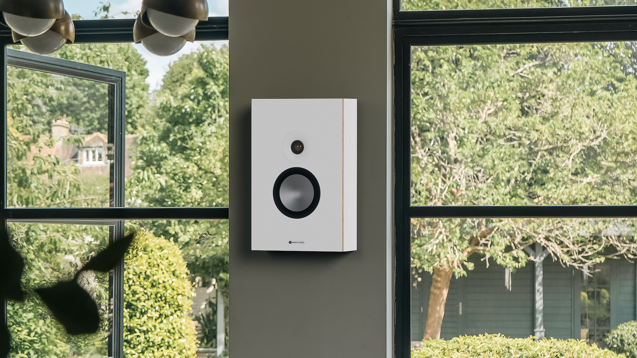 The new Monitor Audio Bronze Series 7G wall-mounted speaker on a wall between two large windows opening onto a bright green garden