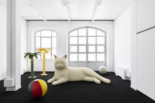 Inside a white room with two beach balls, a yellow and green beach tree and a large white cat