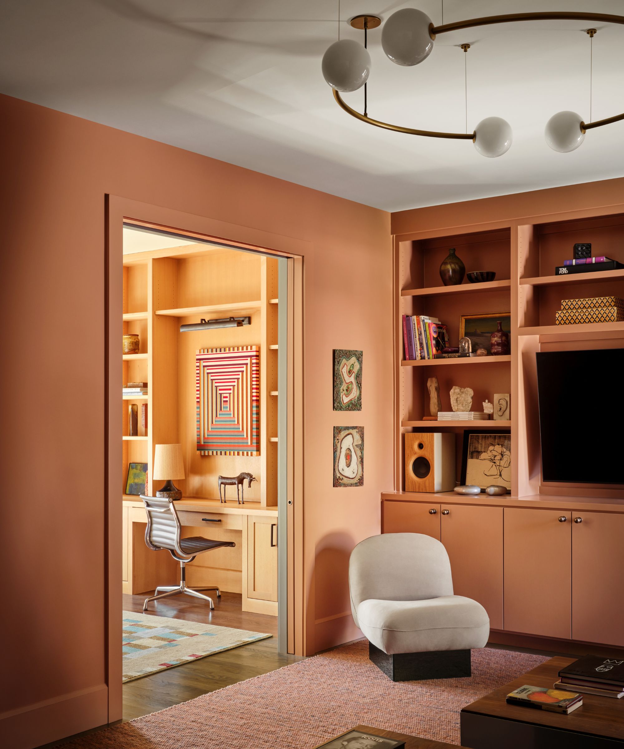 Salmon living room with large pendant light, boucle armchairm, built-in bookshelves and tv looking into orange study
