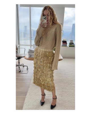 Olympia wears a gold sweater, gold fringe skirt, and black pointed toe heels.