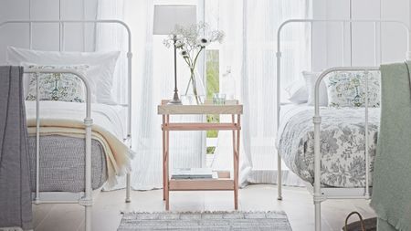 A bright white bedroom with white beds, a large window with sheer curtains, light wooden flooring, a grey rug, a white lamp and a glass vase filled with flowers.